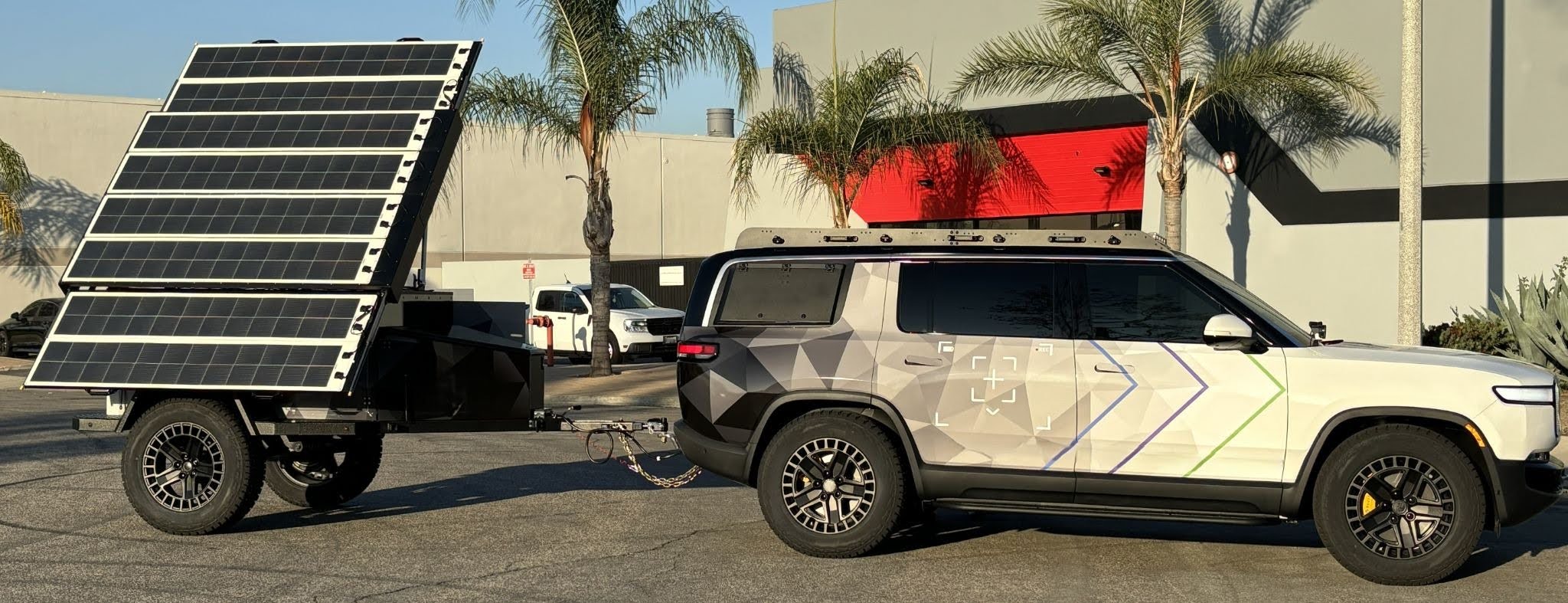 An all-electric SUV towing a deployable solar power station trailer with its panel array raised, parked outside an industrial building.